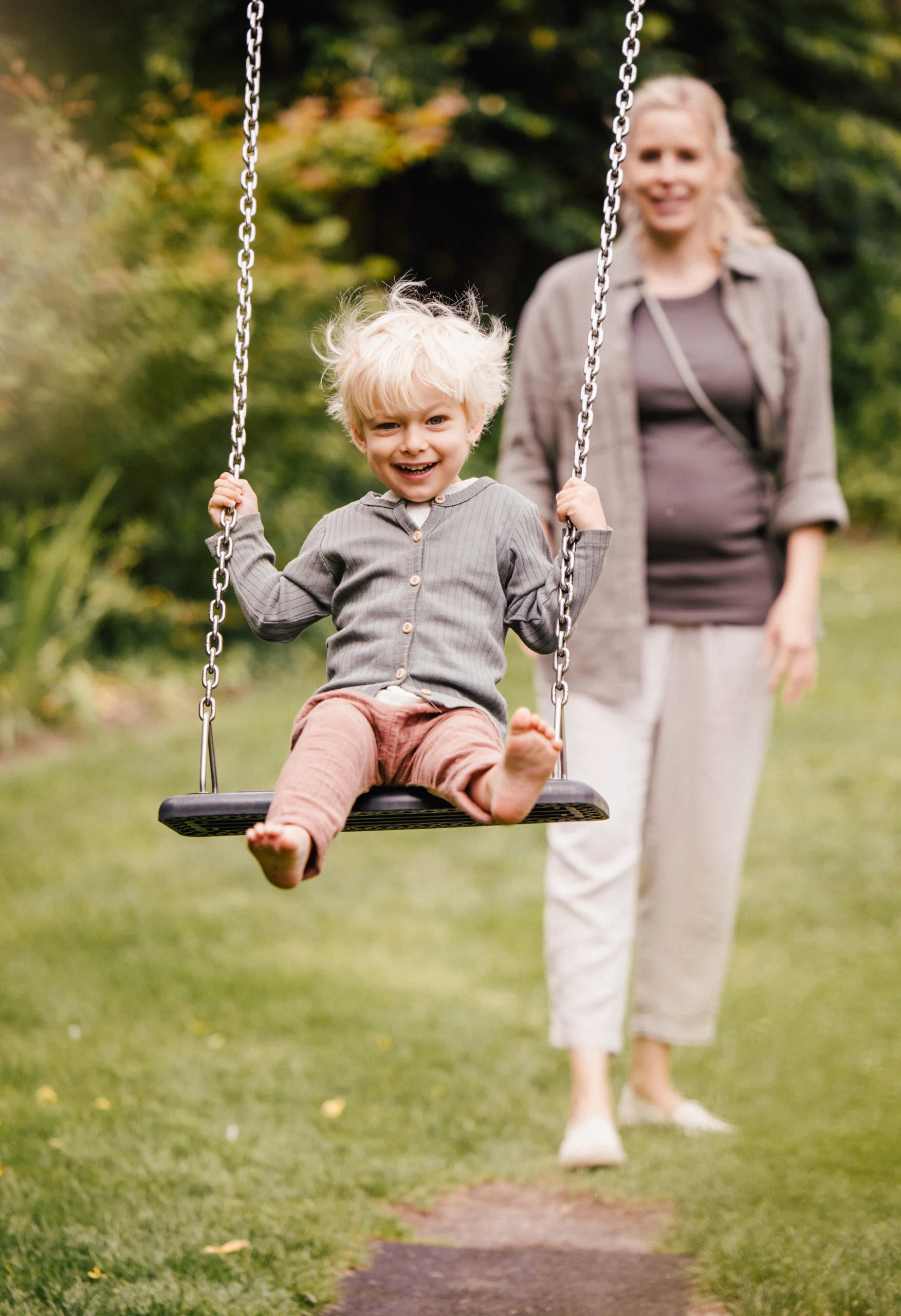 Lachendes Kind auf der Schaukel, während die Mutter im Garten anschubst.