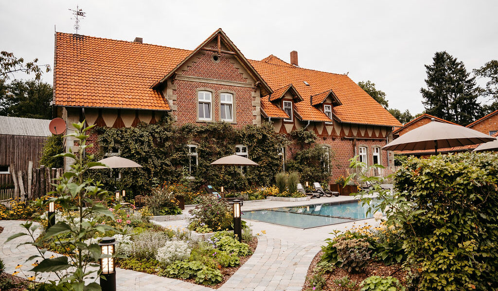 Der Außenpool umgeben von vielen Blumenbeeten, im Hintergrund das rustikale Landhotel