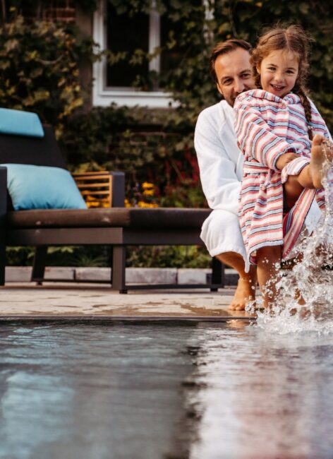 Kleines Mädchen steht mit ihrem Vater am Beckenrand im Kinderhotel mit Schwimmbad und planscht