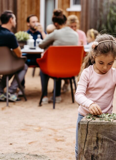 Kleines Mädchen spielt an einem Holzblock mit ihrer Familie im Hintergrund im Hotel mit Kinderbetreuung