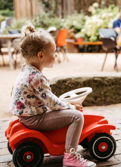 Kleines Mädchen sitzt auf einem roten Bobby-Car im Hotel mit Kinderbetreuung