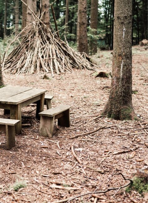 Holzbank  und -tisch im Wald mit einer Holzhütte aus Stöcken im Hintergrund