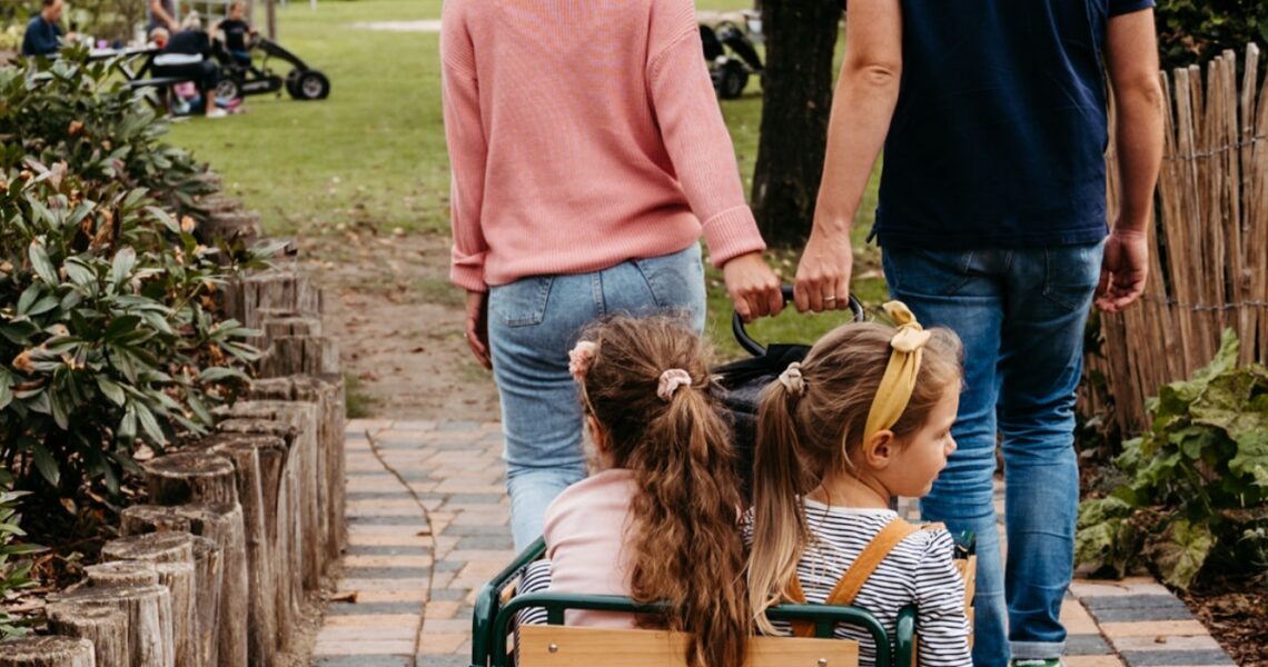 Zwei junge Mädchen sitzen in einem Bollerwagen und werden von den Eltern gezogen.