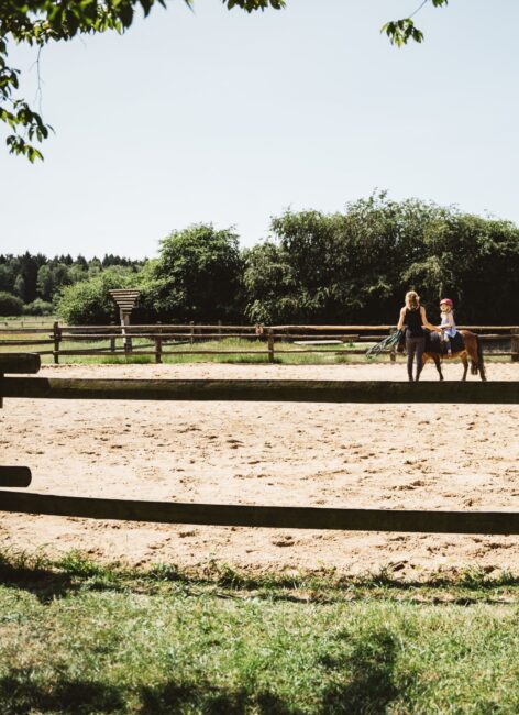 Reitlehrerin steht mit kleinem Mädchen, das auf einem Pony sitzt auf dem Reitplatz im Hotel mit Kinderbetreuung