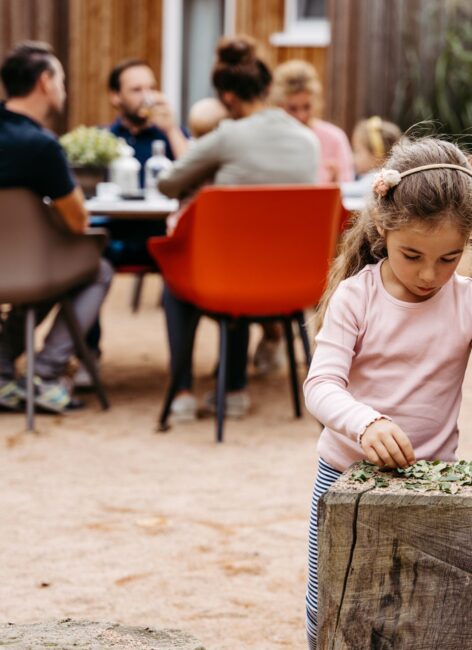Kleines Mädchen spielt an einem großen Holzklotz, während ihre Familie zusammen am Tisch im Familienhotel Niedersachsen sitzt