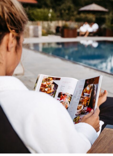 Frau sitzt mit Bademantel am Pool des Familienhotels in Niedersachsen und liest das Menü