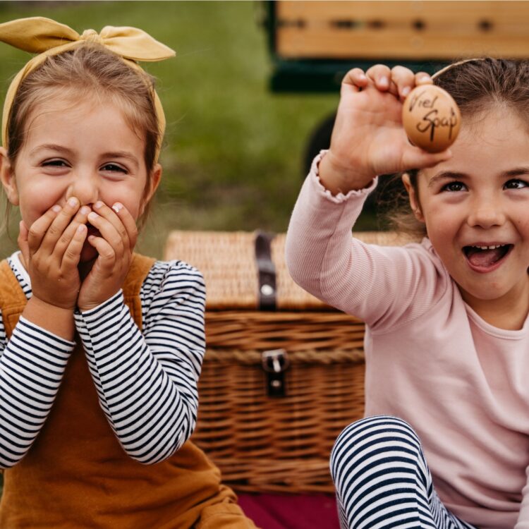 Zwei lachende Mädchen sitzen auf einer Wiese und halten ein Hühnerei in die Kamera