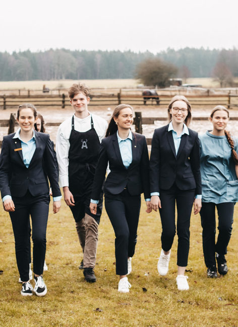 Gruppe von jungen Leuten in Anzug, Kochjacke und Blazer laufen stolz und lächelnd auf dem Hof umher.