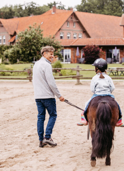Opa führt seine Enkeltochter auf einem Reitplatz auf einem Pony.