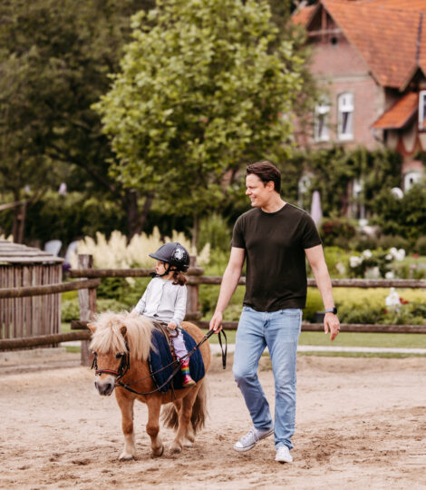 Ein Vater führt sein Kind auf dem Pony über den Reitplatz.