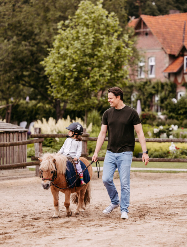 Ein Vater führt sein Kind auf dem Pony über den Reitplatz.