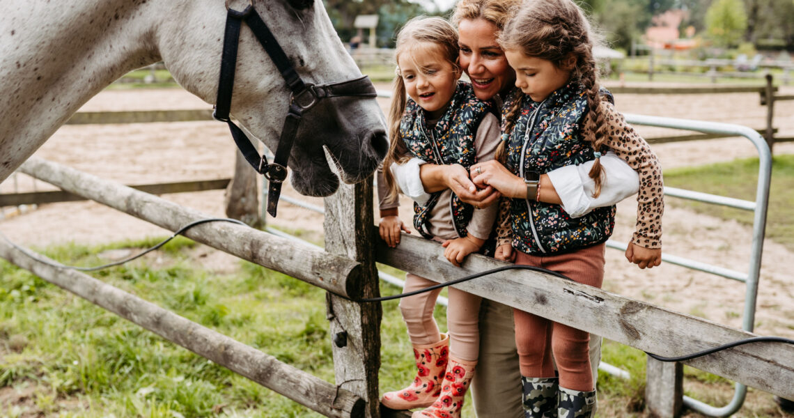 Mutter steht mit ihrem Töchtern am Zaun des Pferdestalls und streichelt ein Pferd mit Familienhotel mit Bauernhof