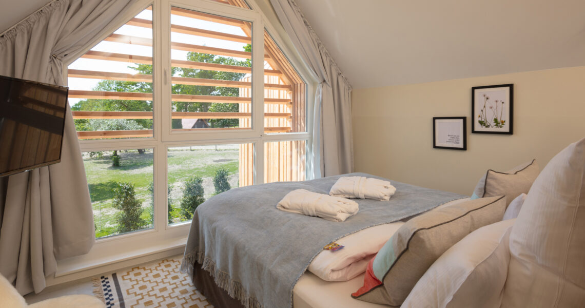 Blick in das Zimmer mit gemütlichem Bett, auf dem Bademäntel liegen , Fernseher und großen Fenstern