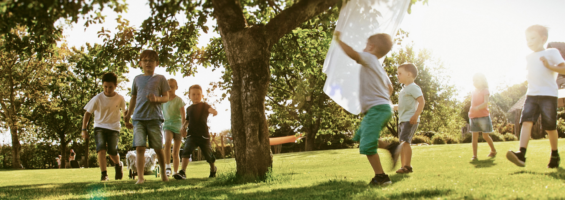 Sieben kleine Kinder spielen auf einer grünen Wiese unter einem Baum beim Landhaus Averbeck