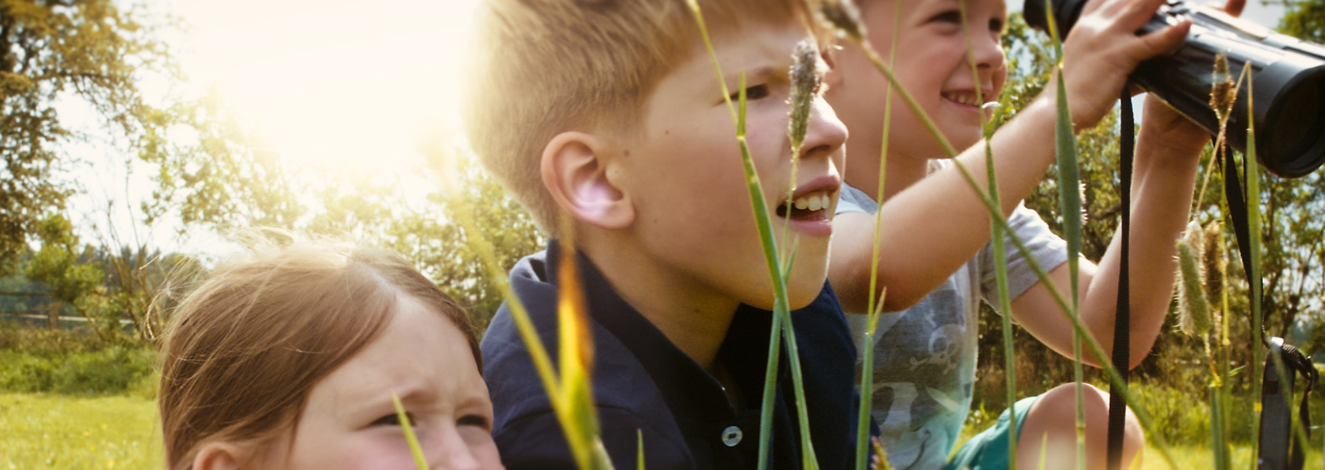 Zwei Jungs und ein Mädchen sitzen mit einem Fernglas auf der Wiese beim all inclusive Familienurlaub