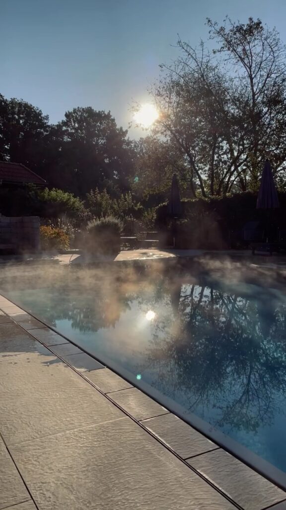 Früh morgens am Pool, der Himmel spiegelt sich im flachen Wasser, Ruhe liegt über dem Hof, die erste…